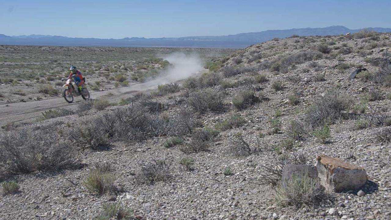 The desert terrain is harsh, as evidenced by the foreground, and the racers are flying through it with incredible speed to stay ahead of their competitors.